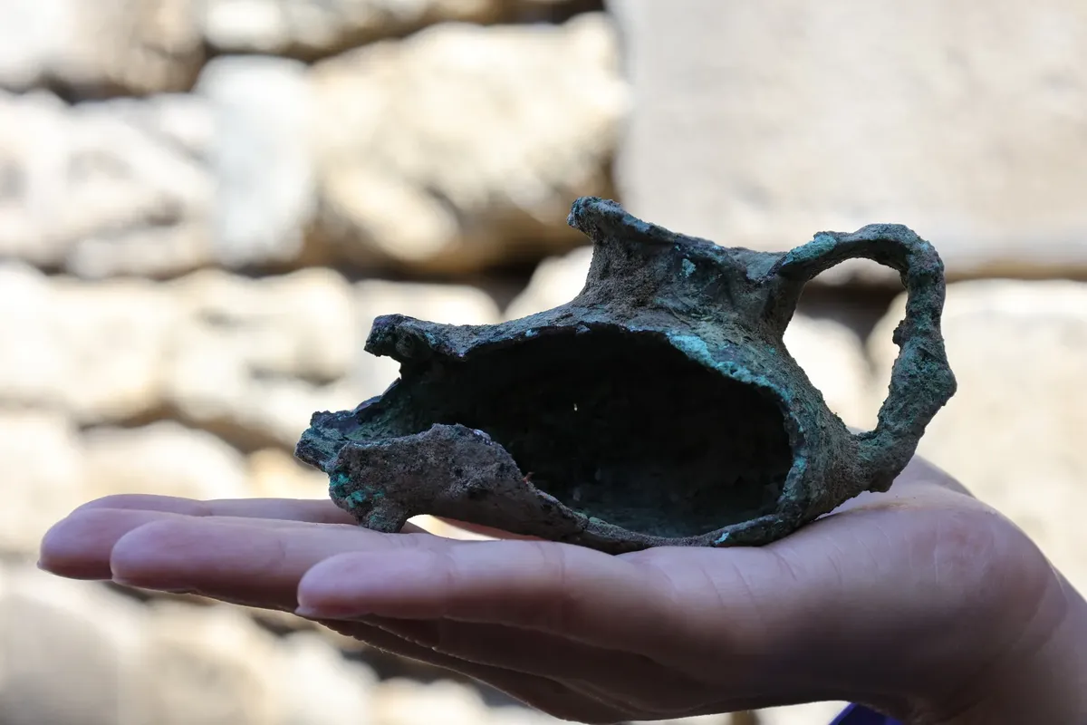 Close-up of a hand holding an ancient bronze artifact against a stone background.
